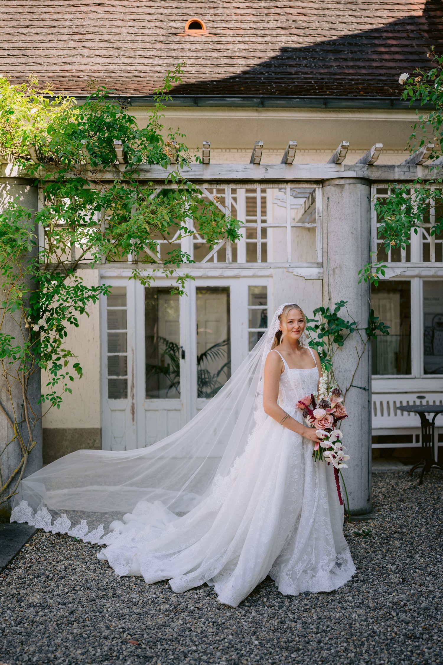 Bride in a lace ball gown with cathedral veil holds a bouquet outside an elegant venue with lush greenery.