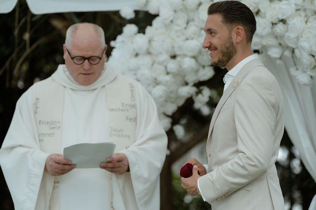A wedding ceremony takes place with an officiating priest in white robes reading from notes beside a groom in white suit.