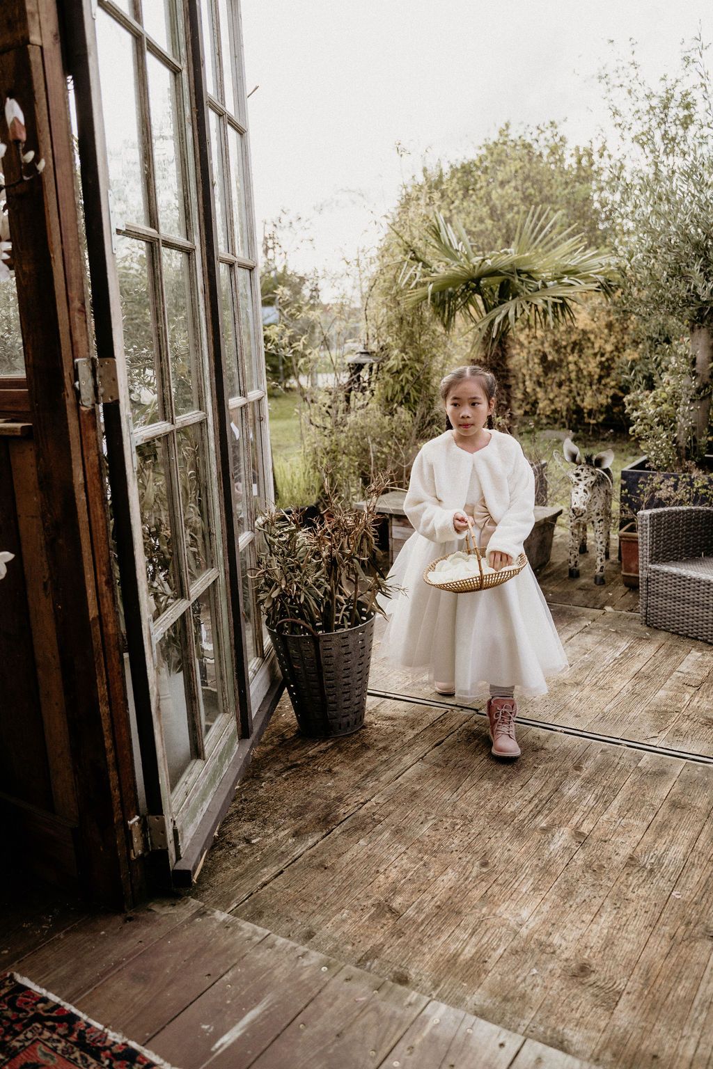 Child in white dress sits on rustic wooden deck surrounded by potted plants and glass windows creating cozy atmosphere.
