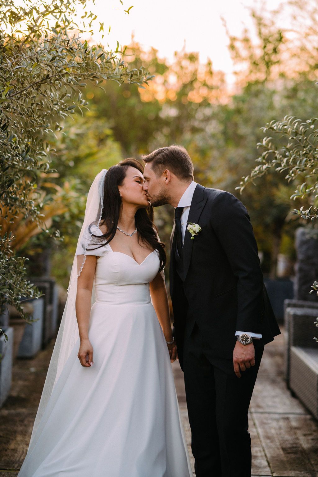 A romantic wedding photograph capturing a gentle kiss between newlyweds in a garden setting at sunset.