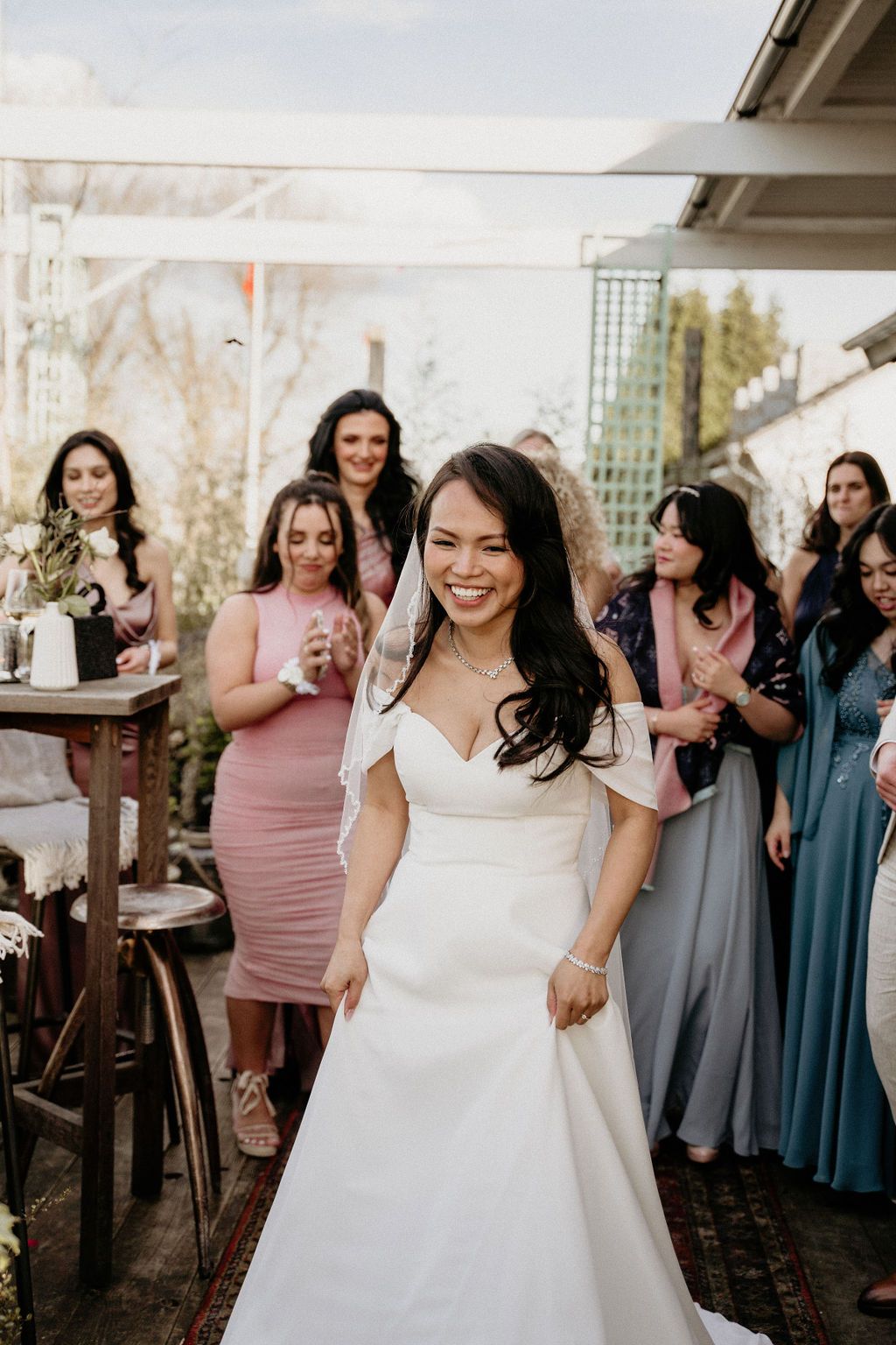 A bride in a white wedding dress celebrates joyfully with guests and wedding party members wearing pink and blue dresses.