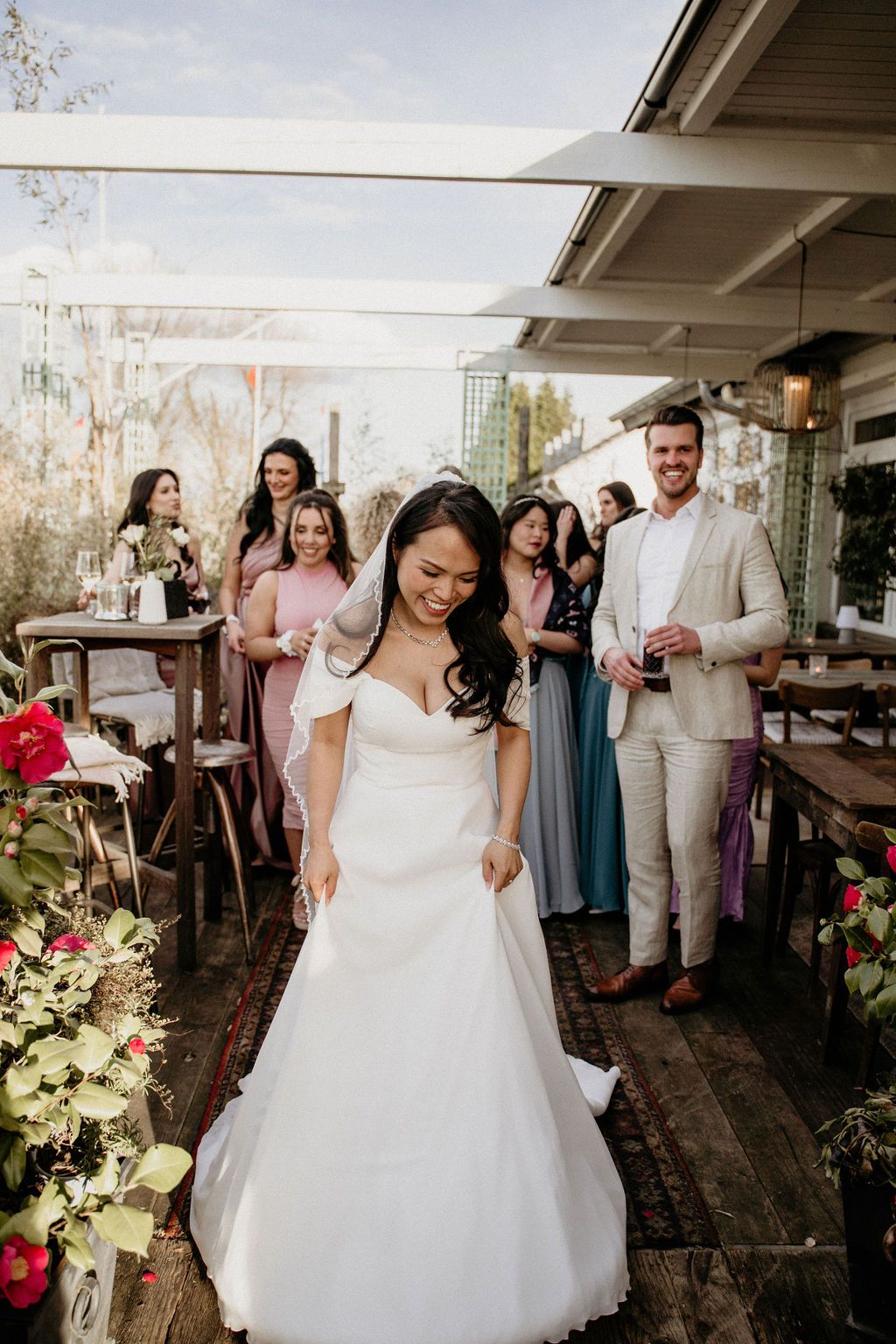 Wedding party gathers on an outdoor patio as bride in white gown stands in focus with guests in pink dresses behind her.