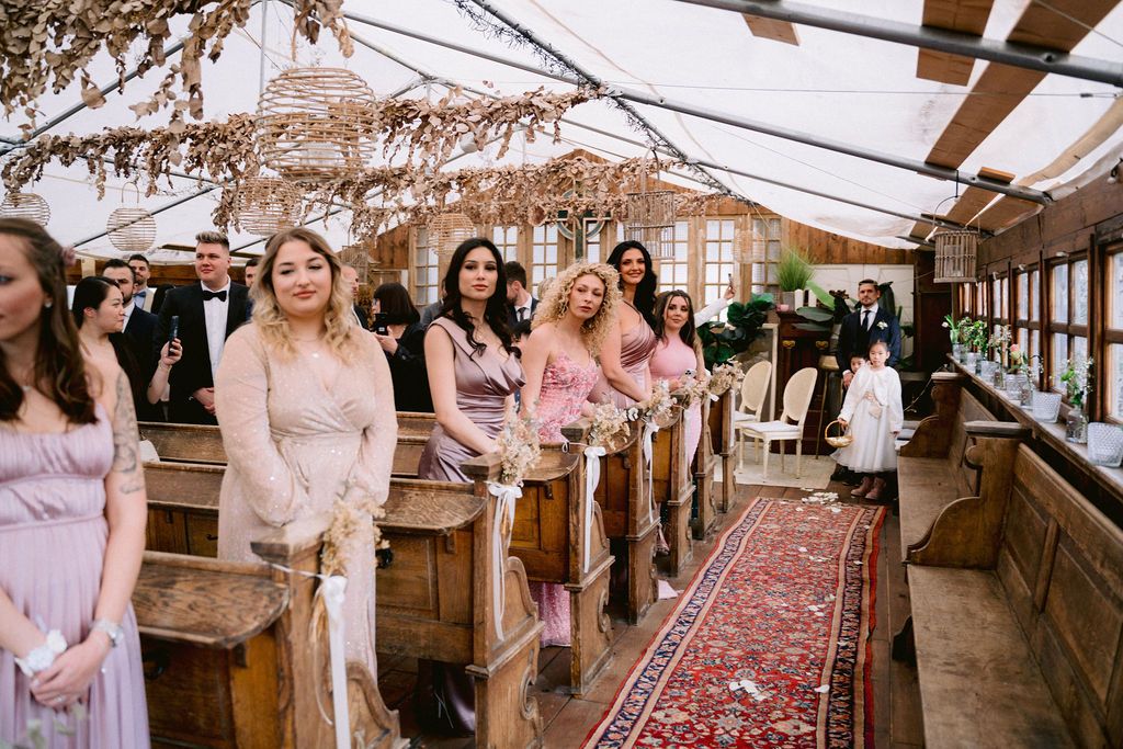 Row of people standing at rustic indoor venue with wooden beams and oriental rugs lining the floor for wedding celebration.