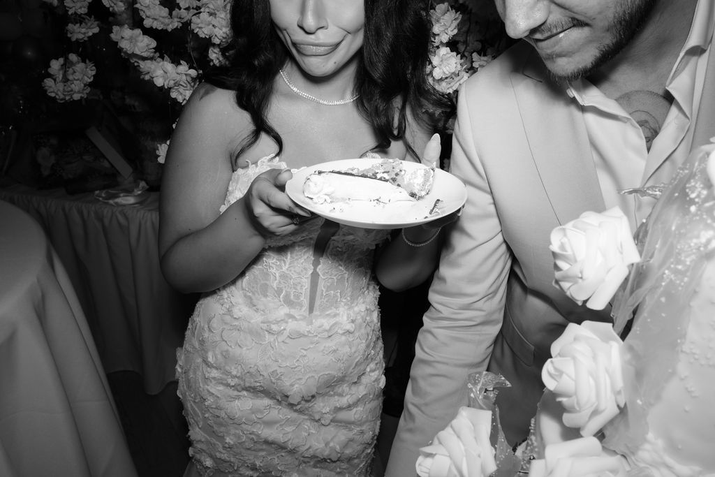 A black and white photo captures a couple sharing dessert at what appears to be a wedding reception.