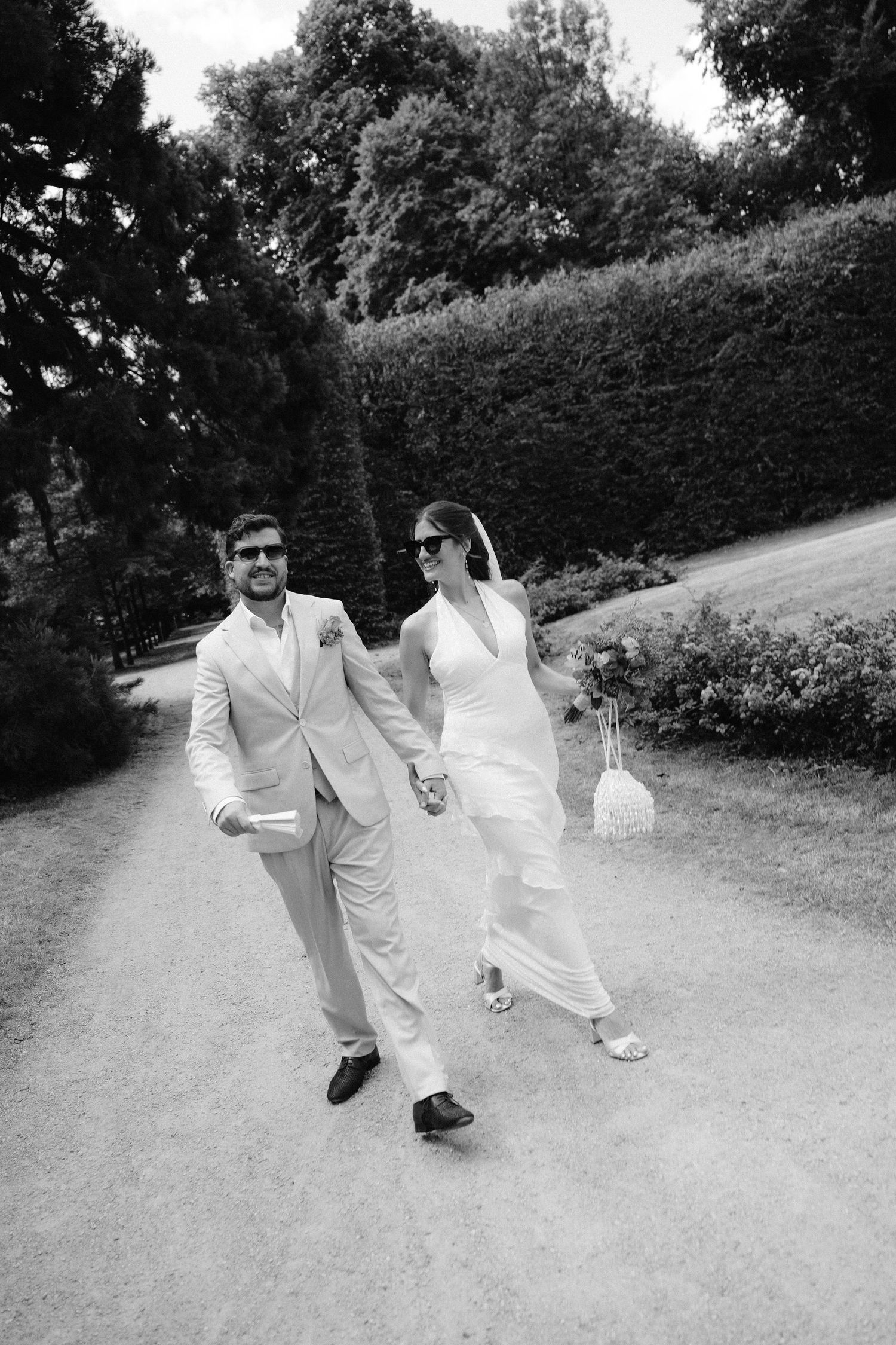 Black and white photo of a stylish bride and groom holding hands, walking together on a garden path on their wedding day.