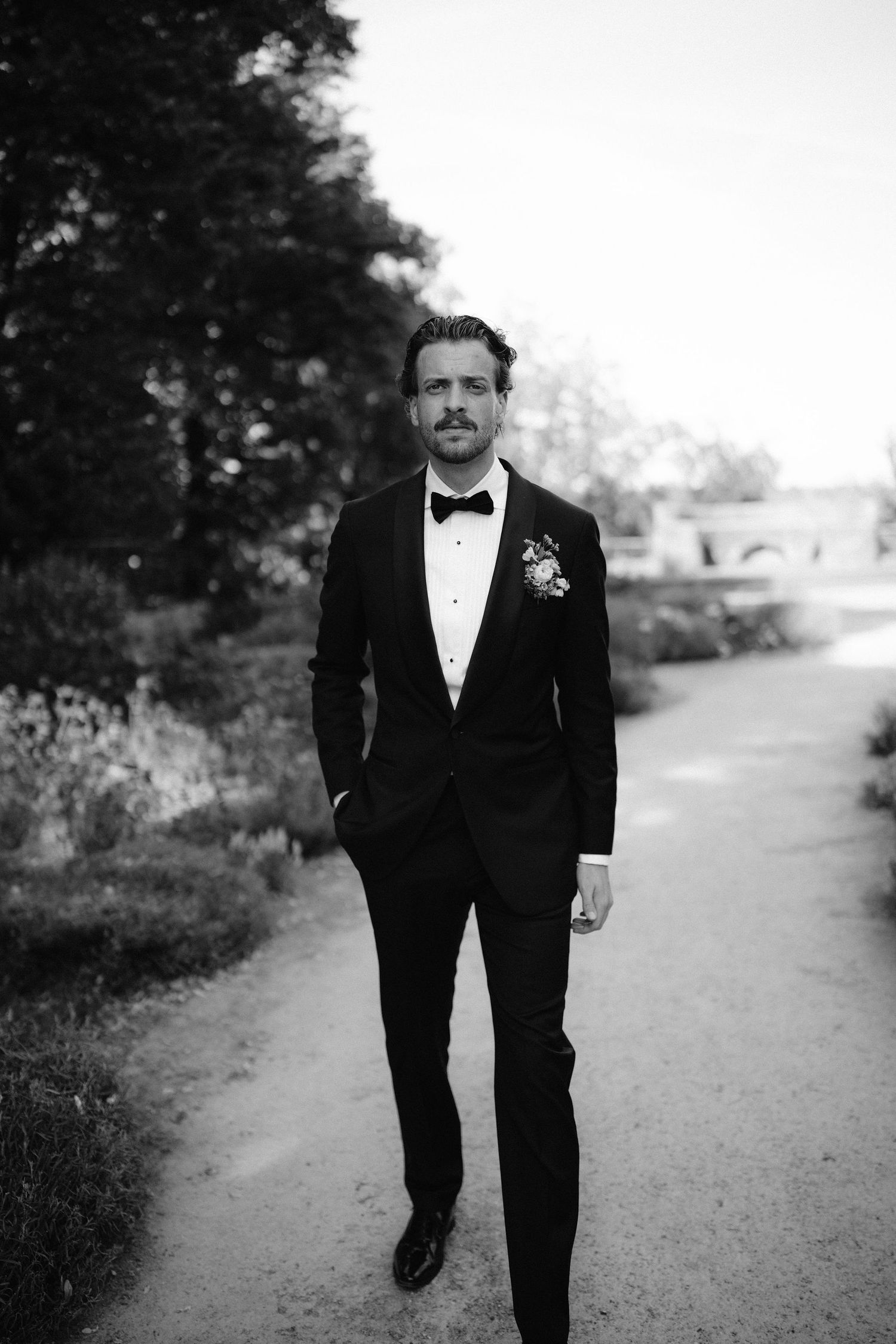 Elegant groom in black tuxedo with bow tie and boutonniere walks garden path in black and white photo.