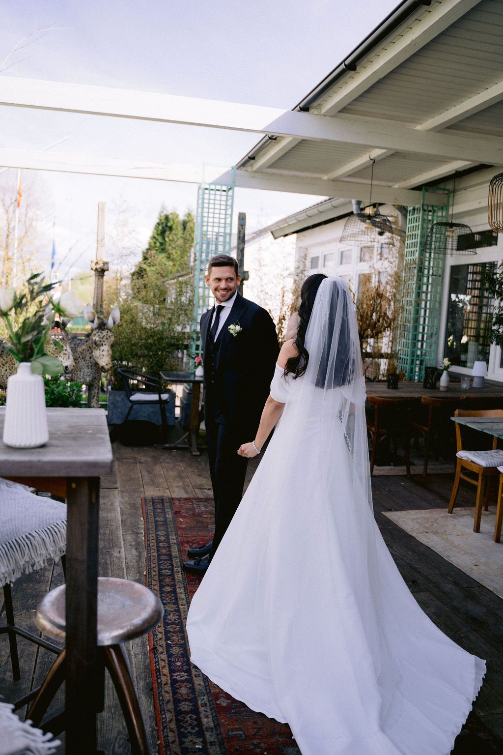 A bride in a flowing white gown with veil stands on an outdoor terrace with decorative rugs and seating arrangements.