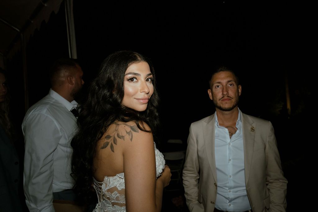 A couple at a dimly lit evening event, with the woman wearing a strapless dress and the man in a tan blazer.