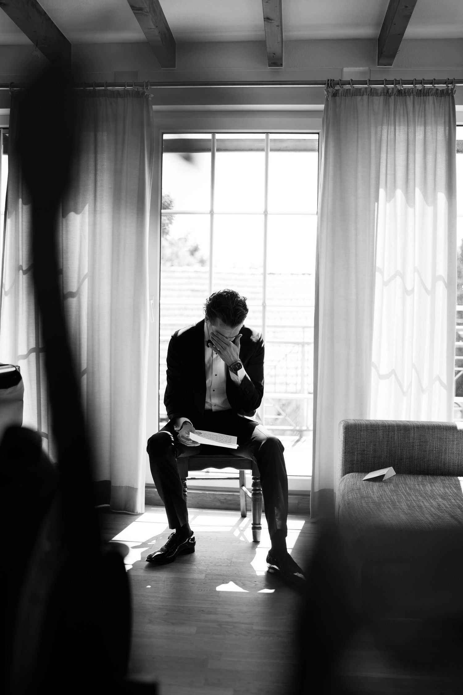 Groom in black suit reads vows while seated by window in black and white wedding preparation photo.