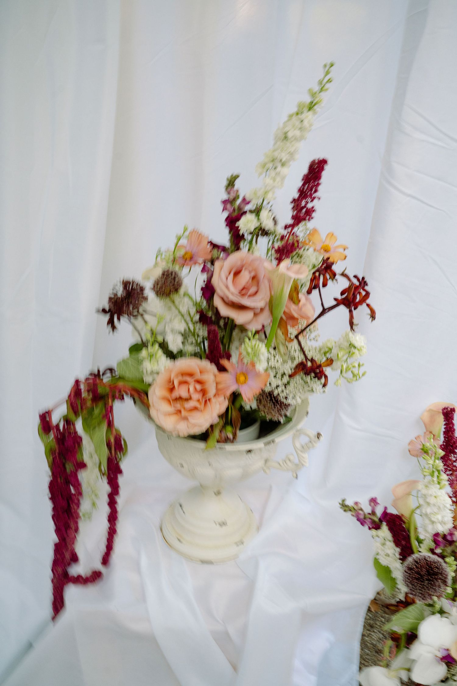 Elegant white urn vase with lush wedding floral arrangement featuring peach roses, burgundy blooms, and wildflowers on draped white fabric.
