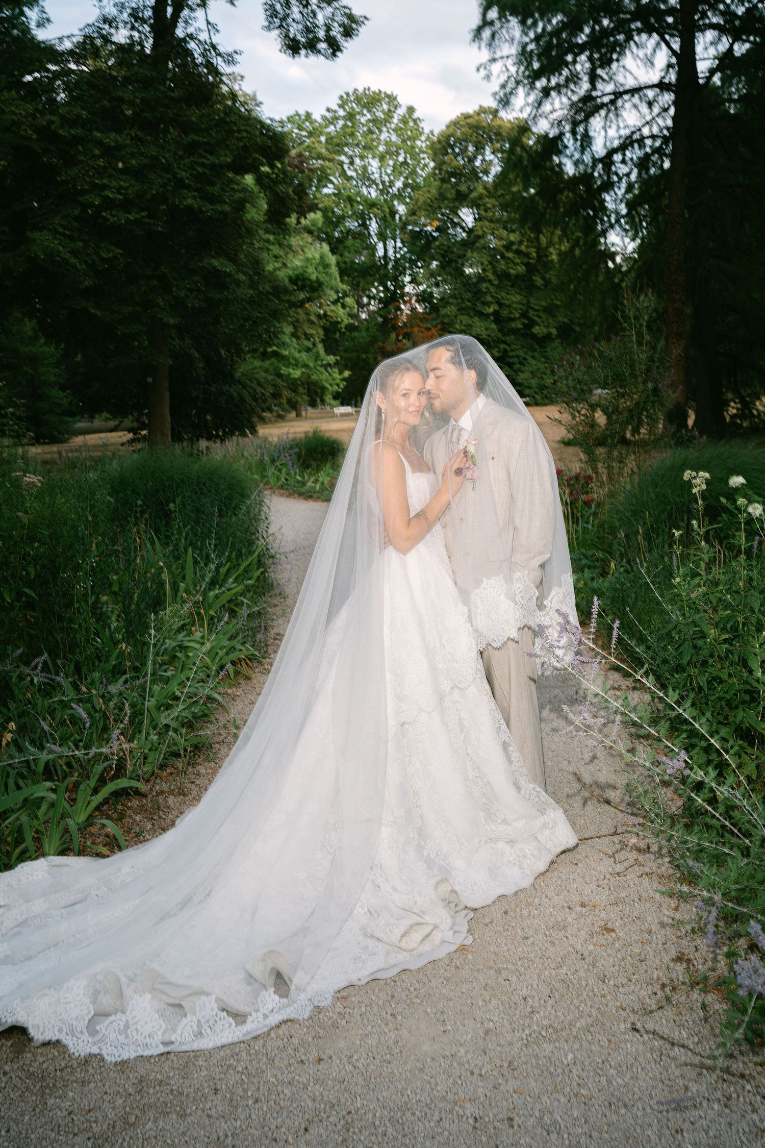 Bride and groom share a romantic moment under a flowing veil in a lush garden setting on their wedding day.