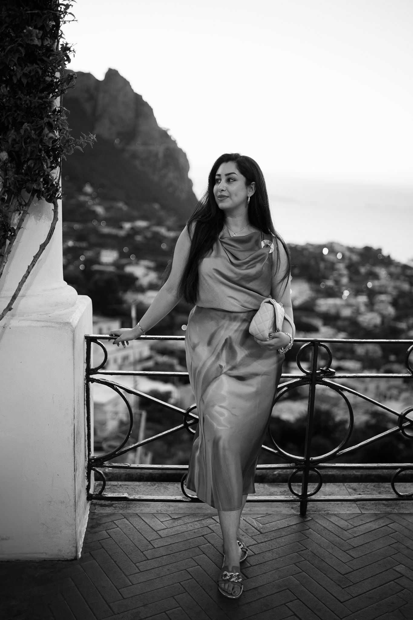 Elegant woman in satin midi dress poses on scenic terrace overlooking mountainous coastal town in black and white.