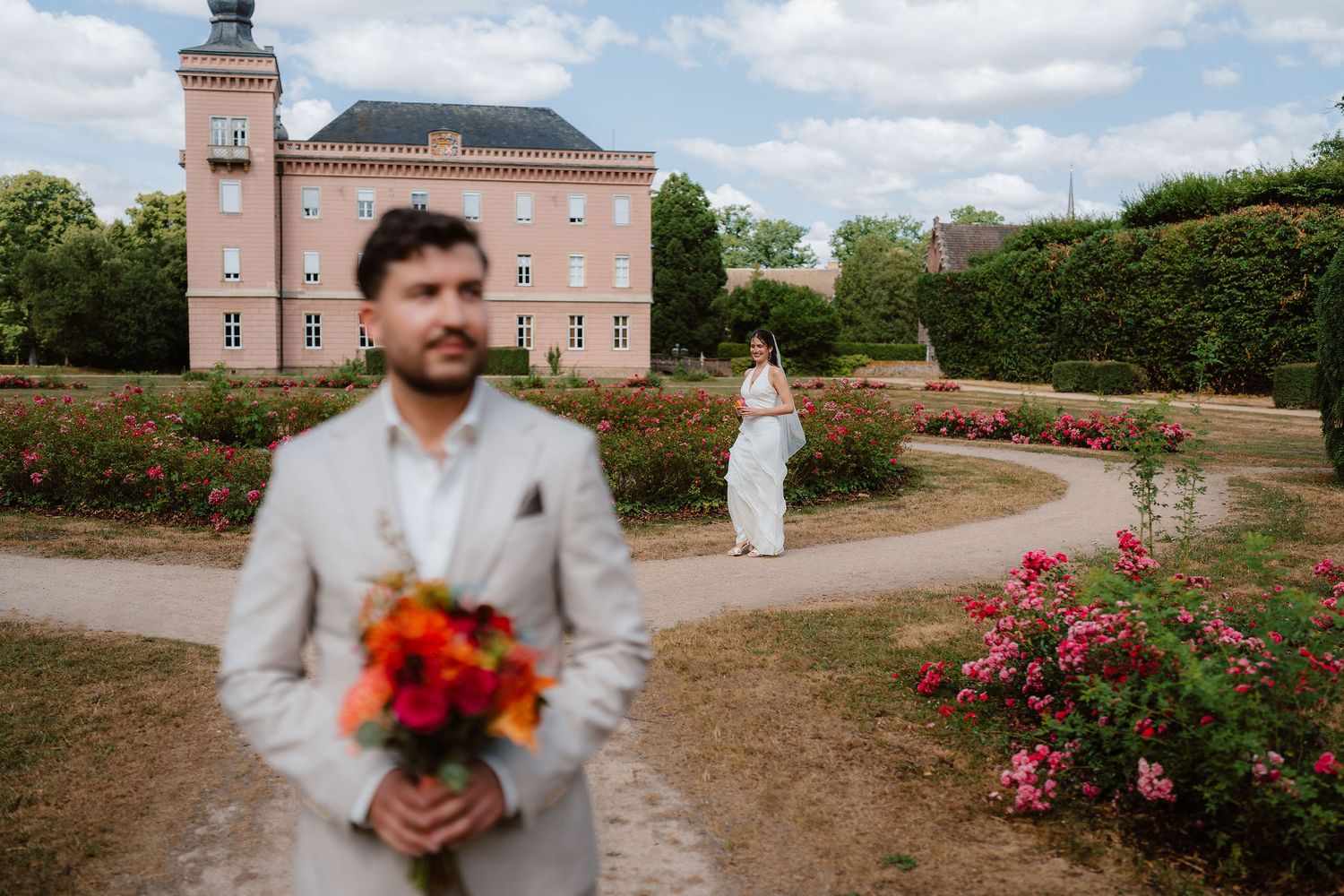 Groom holding colorful bouquet in foreground with bride walking toward pink castle in rose garden.