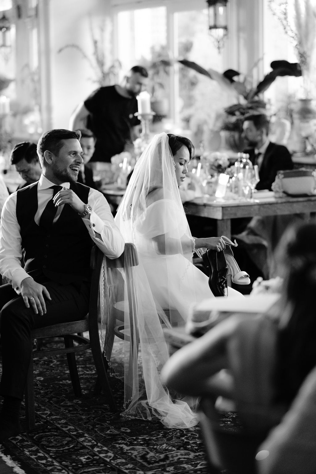 A black and white wedding scene at a reception venue shows a bride in a veil and guests seated at elegantly decorated tables.