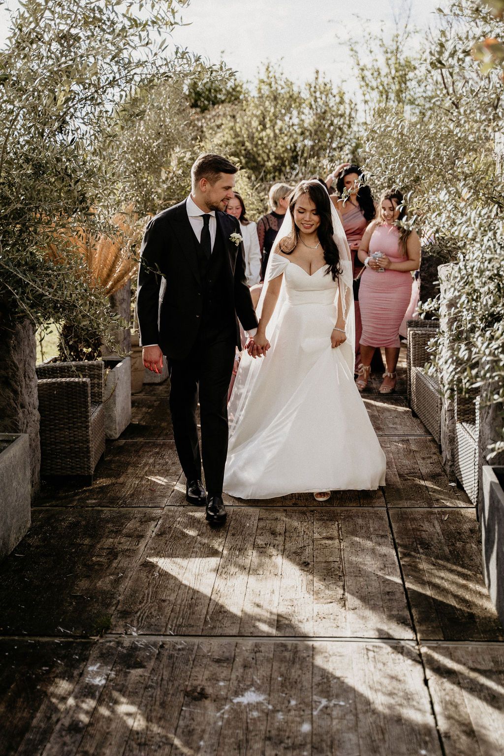 Newlyweds walking together on a rustic wooden pathway surrounded by greenery during an outdoor wedding ceremony.