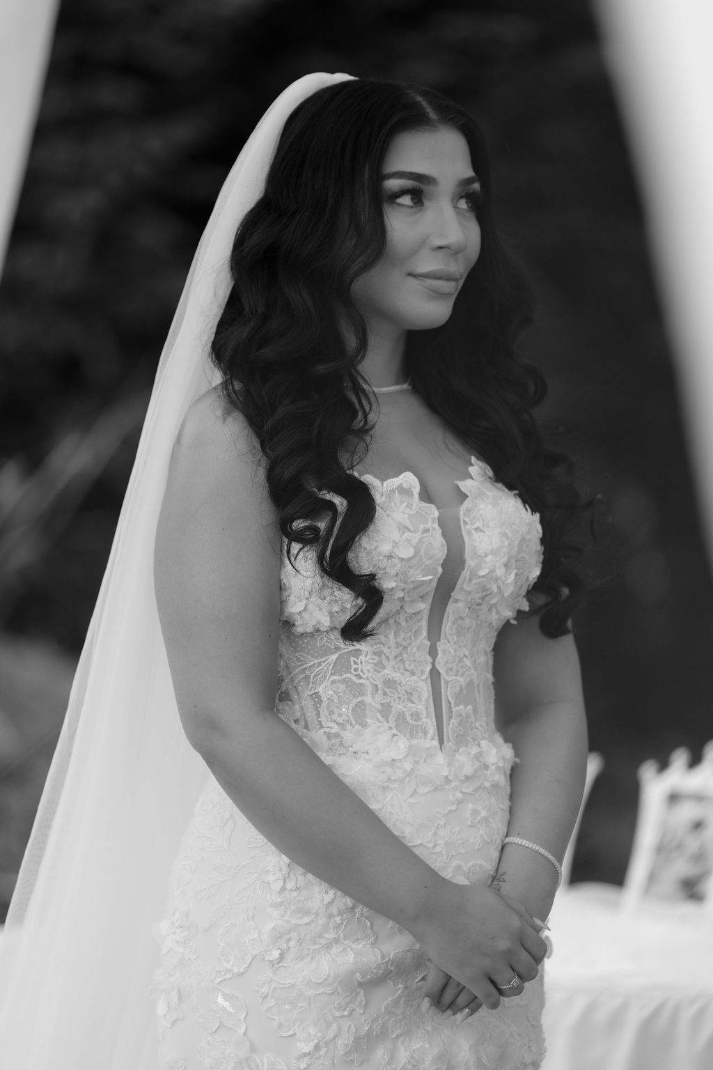 A bride in a white lace wedding dress stands outdoors in elegant bridal portraits taken in black and white.
