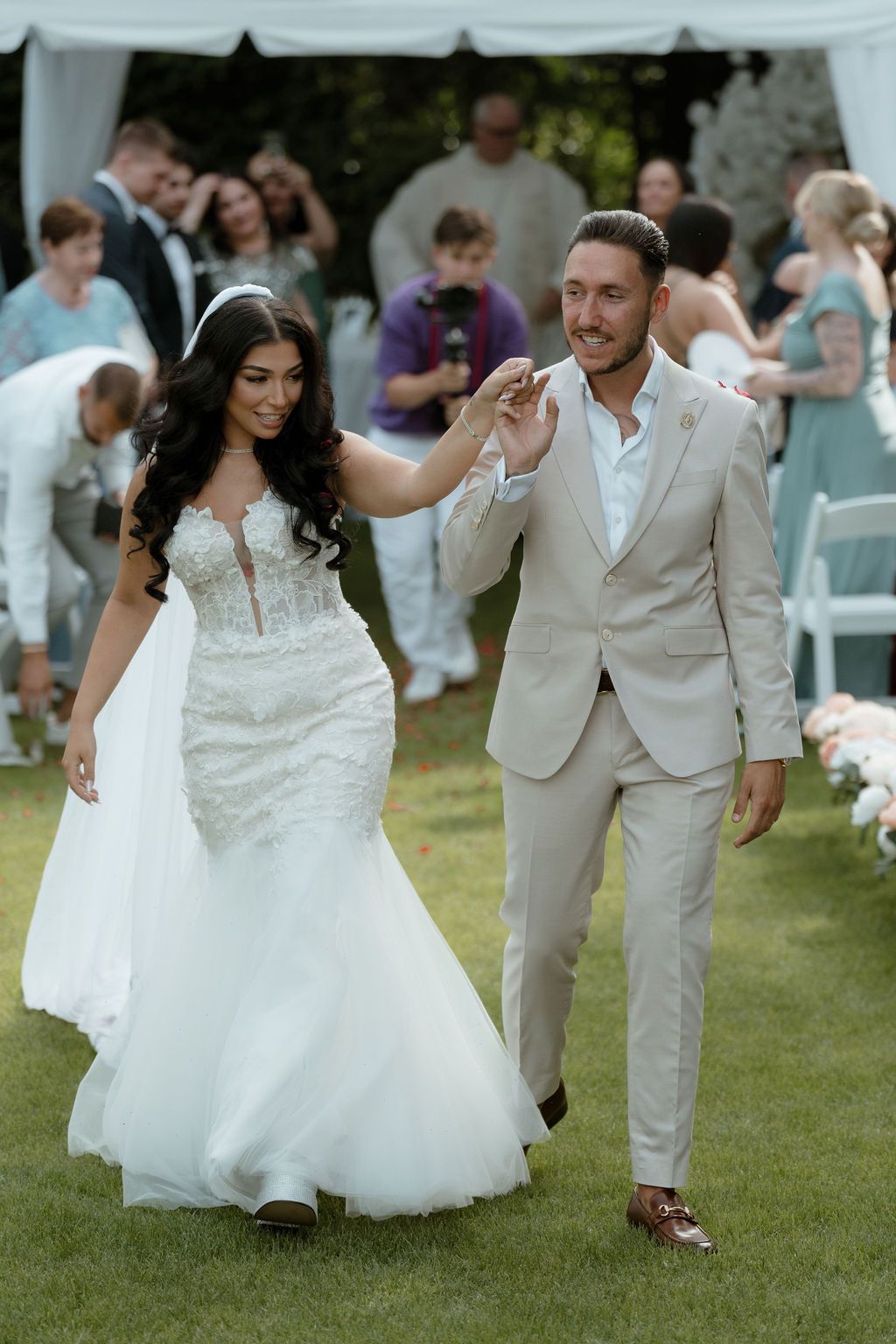 Newlyweds walk hand in hand on a grassy lawn during outdoor garden wedding ceremony with guests in background.