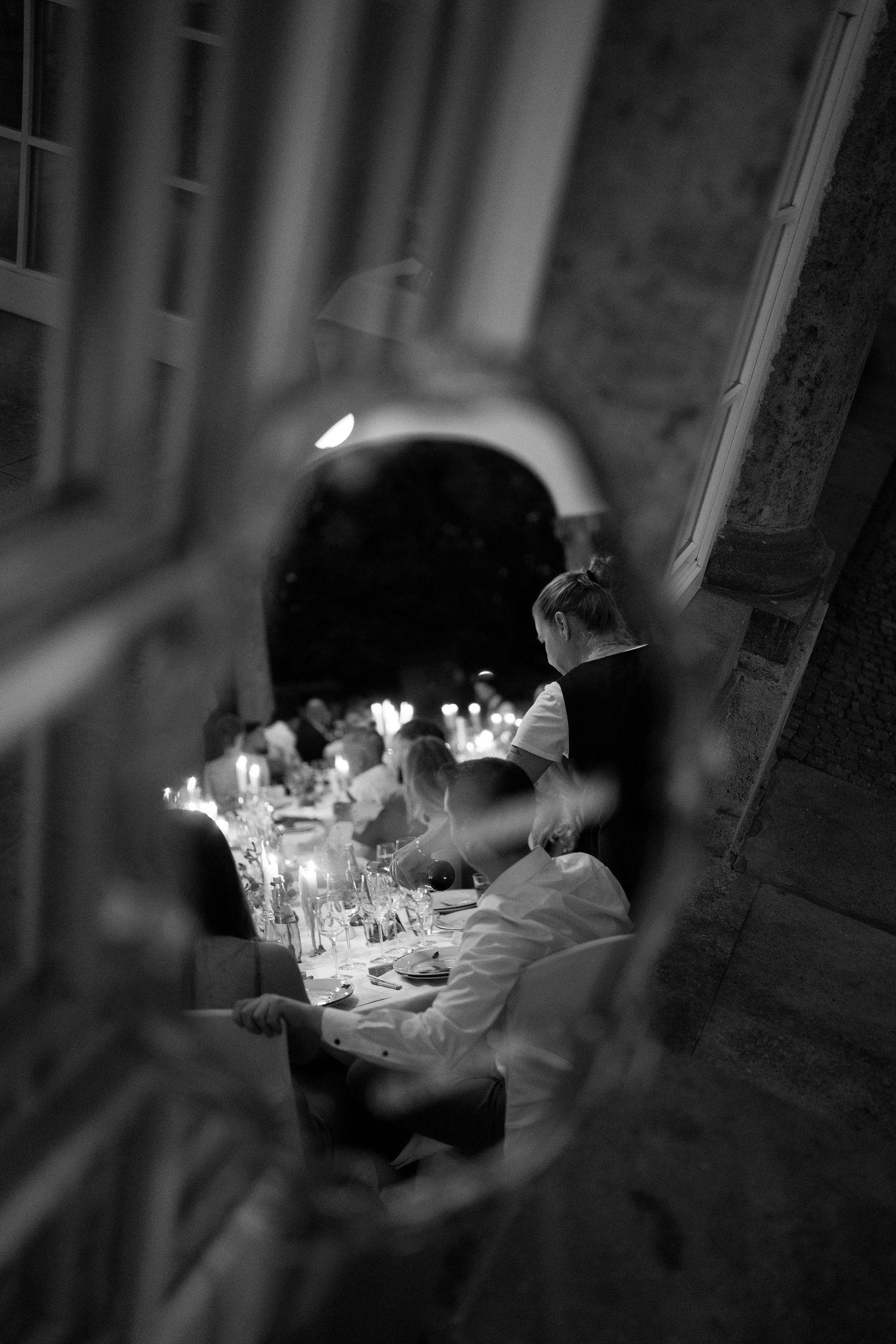 Black and white photo of elegant candlelit dinner party guests seated at a long table viewed through an archway.