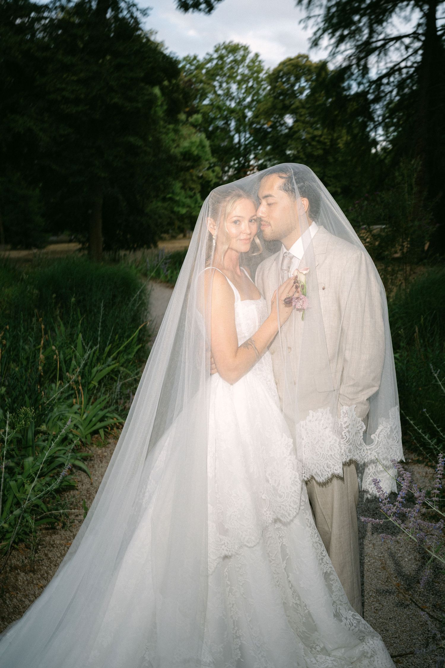 Bride and groom share romantic moment under flowing lace veil in lush garden setting on their wedding day.