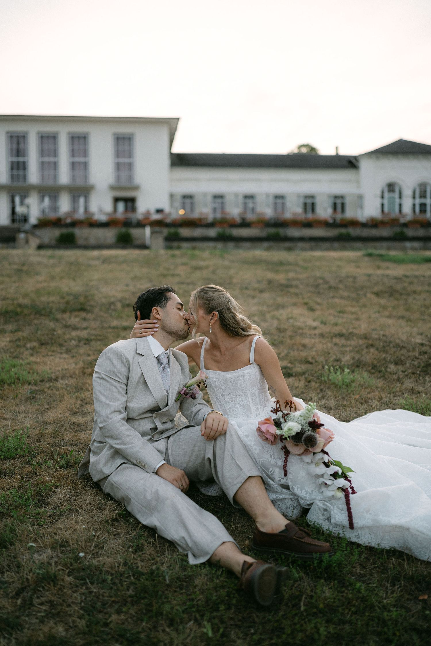 Bride and groom share a romantic kiss while sitting on grass in front of an elegant white venue.