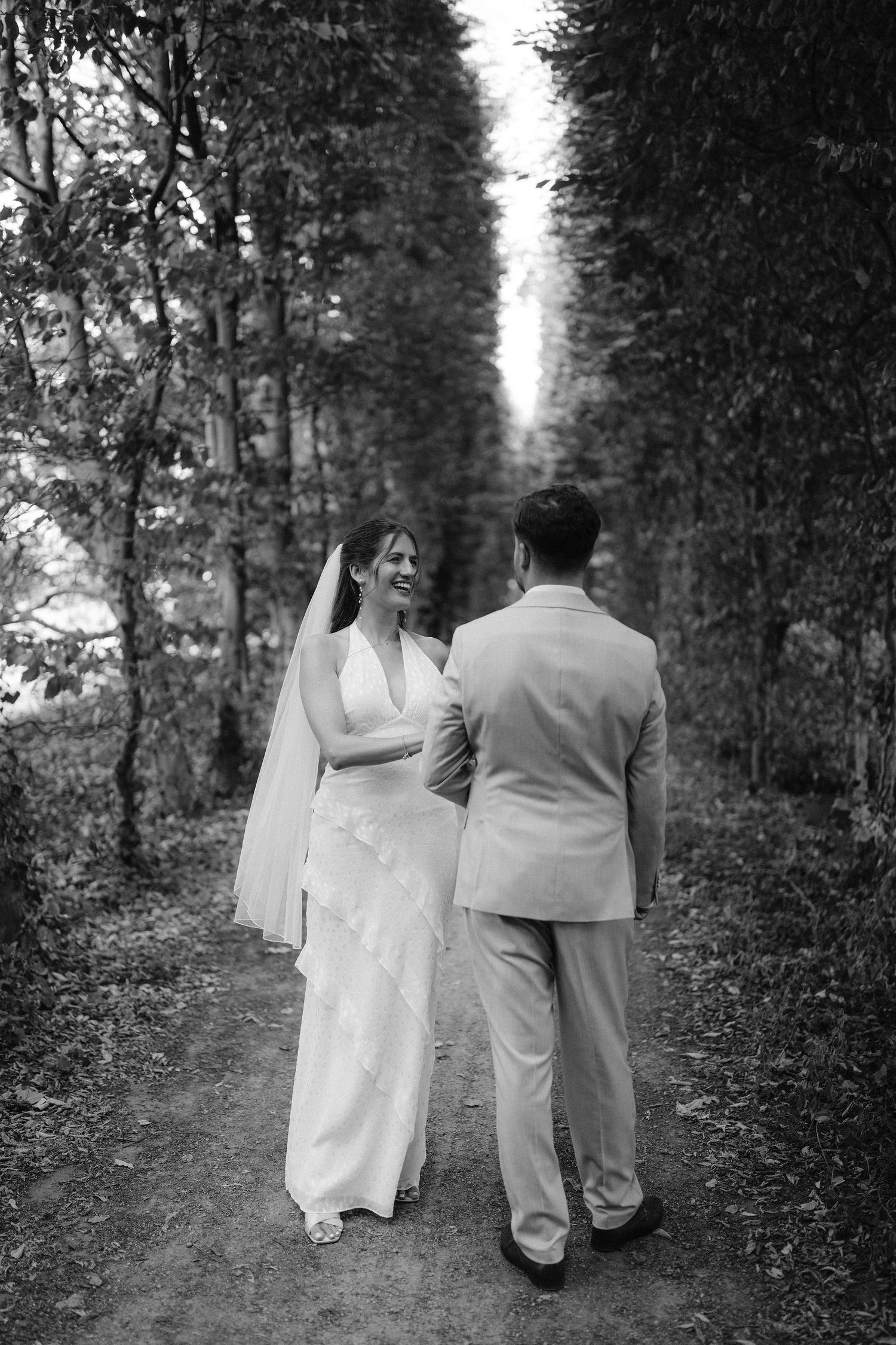 Black and white photo of a smiling bride in a white gown and veil walking hand in hand with a groom in a suit along a tree-lined path.