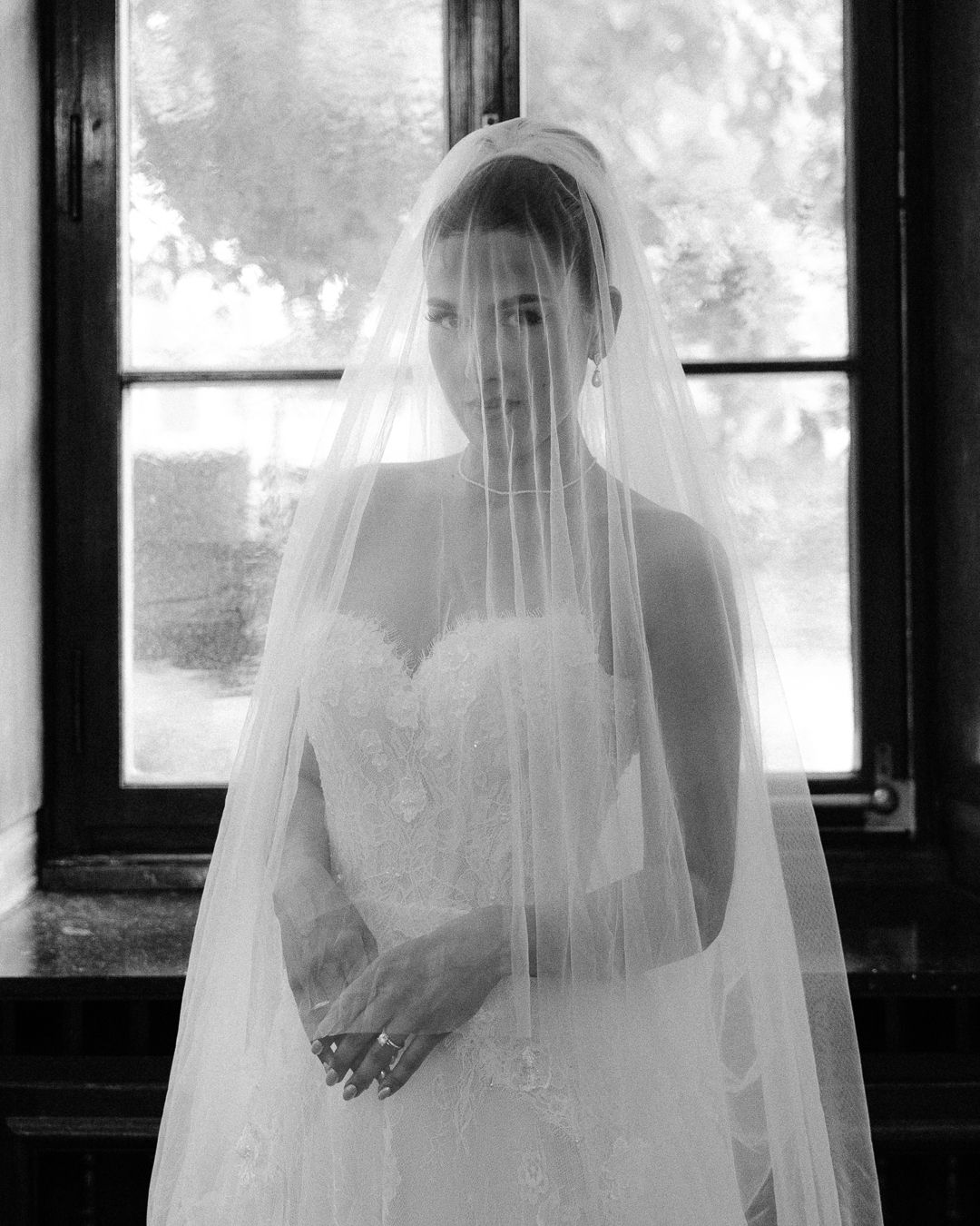 Bride wearing strapless lace wedding gown and long cathedral veil poses gracefully before a window in black and white.