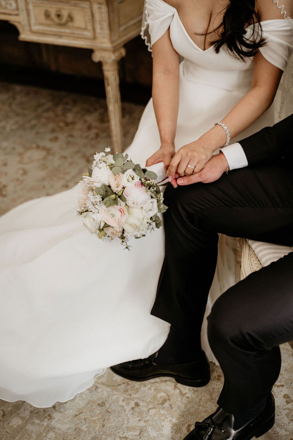 Close-up details of a bride's white wedding dress and groom's black pants with a delicate white floral bouquet between them.