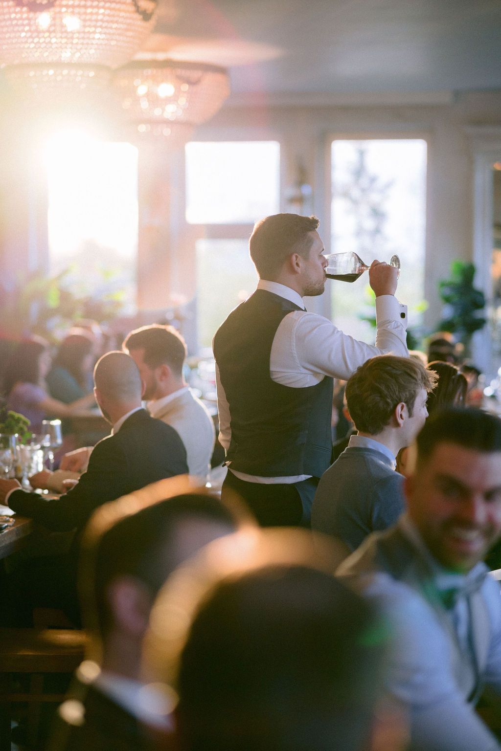 Someone raises a glass in a toast at an elegant wedding reception filled with guests and soft golden light.