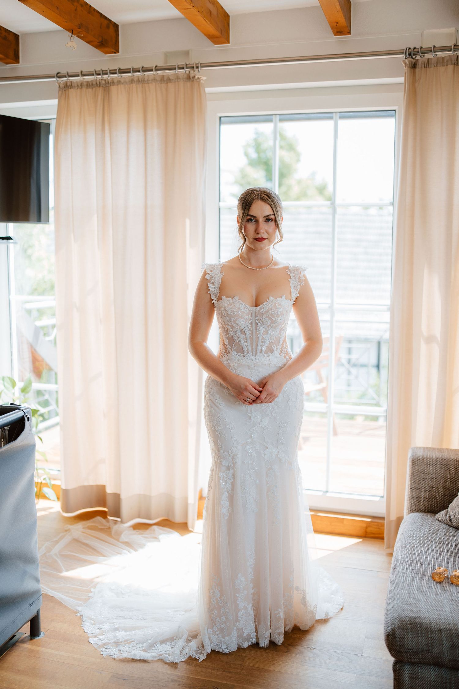 Elegant bride in lace mermaid wedding gown stands before bright window in sunlit room.