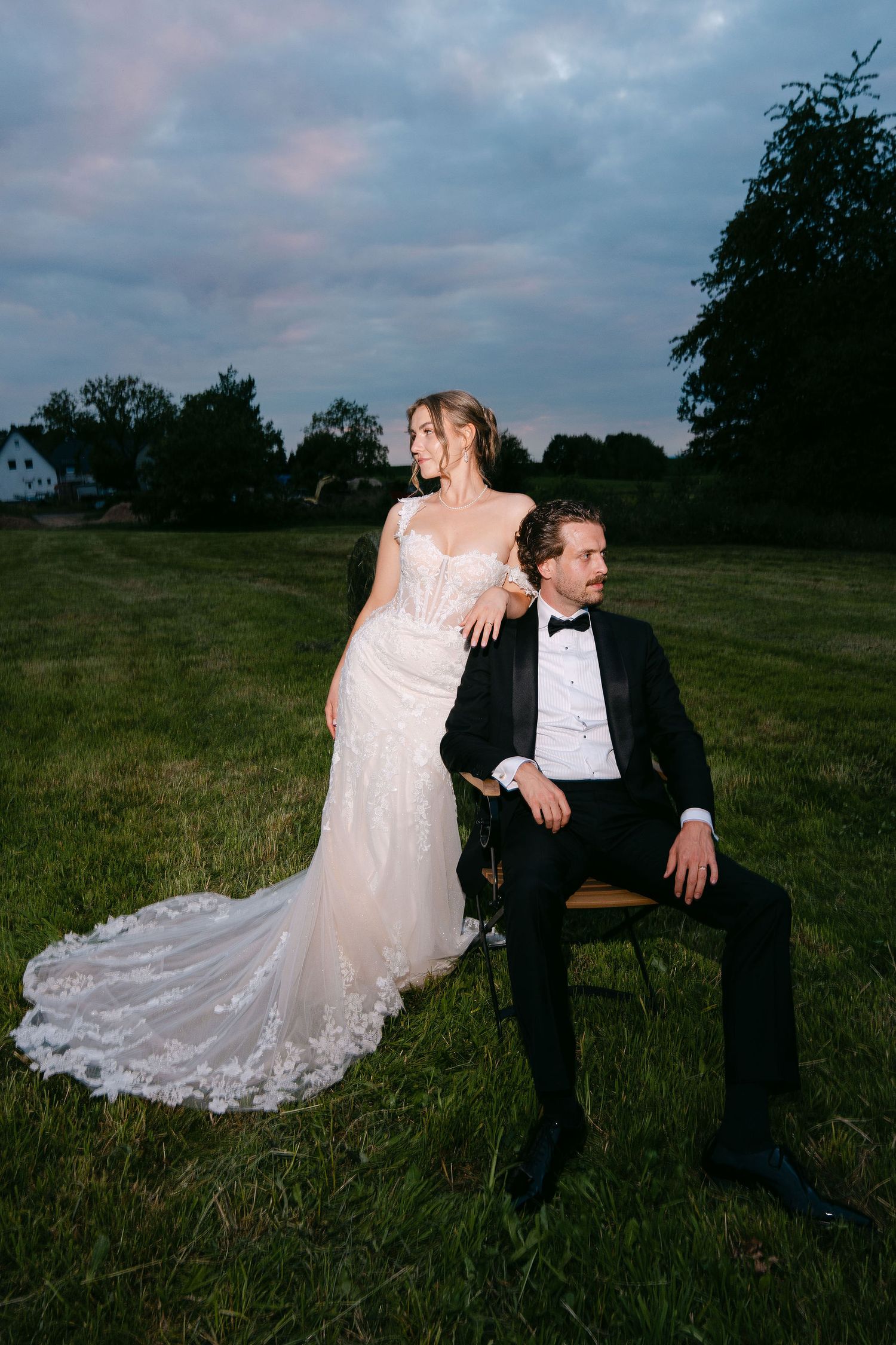 Bride in lace gown stands beside groom in tuxedo seated on chair in green field at dusk.