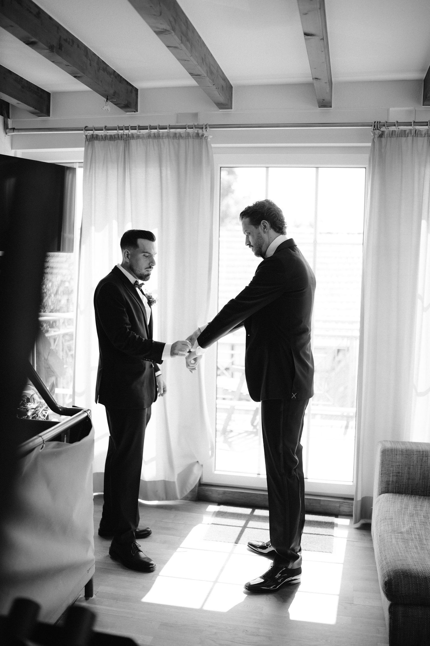 Two men in suits shake hands in a sunlit room with wooden beams, in a classic black and white wedding photo.