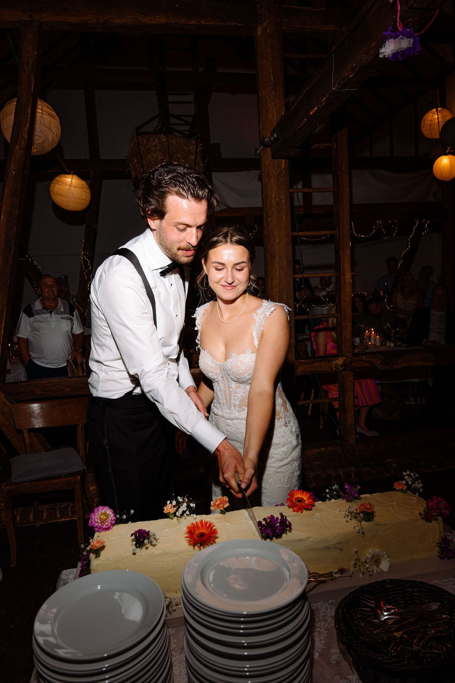 Bride and groom cutting wedding cake together in rustic barn venue with warm lighting and floral decorations.