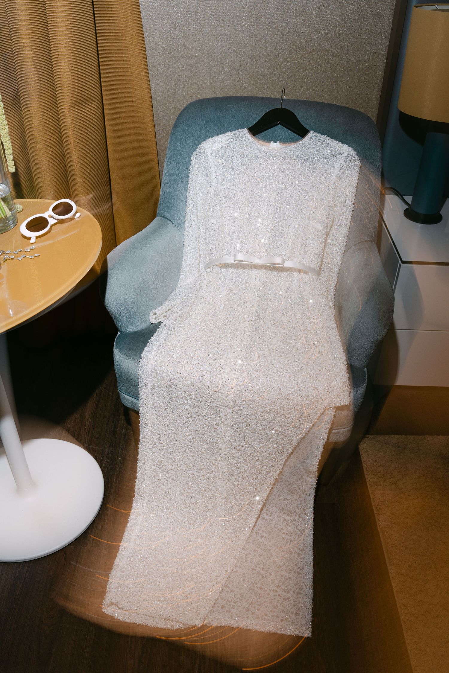 White sequined wedding dress on hanger draped over blue velvet chair with sunglasses on side table nearby.