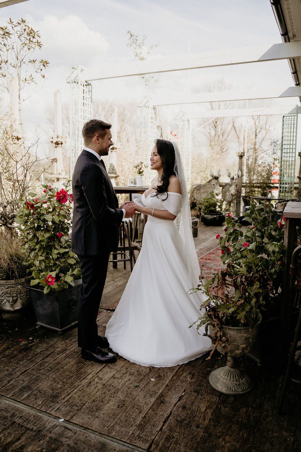 A couple in wedding attire stands on a rustic wood deck surrounded by potted plants and roses.