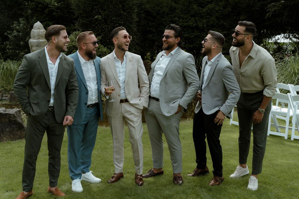 A cheerful wedding party dressed in grey suits gathers on a lawn during an outdoor celebration.