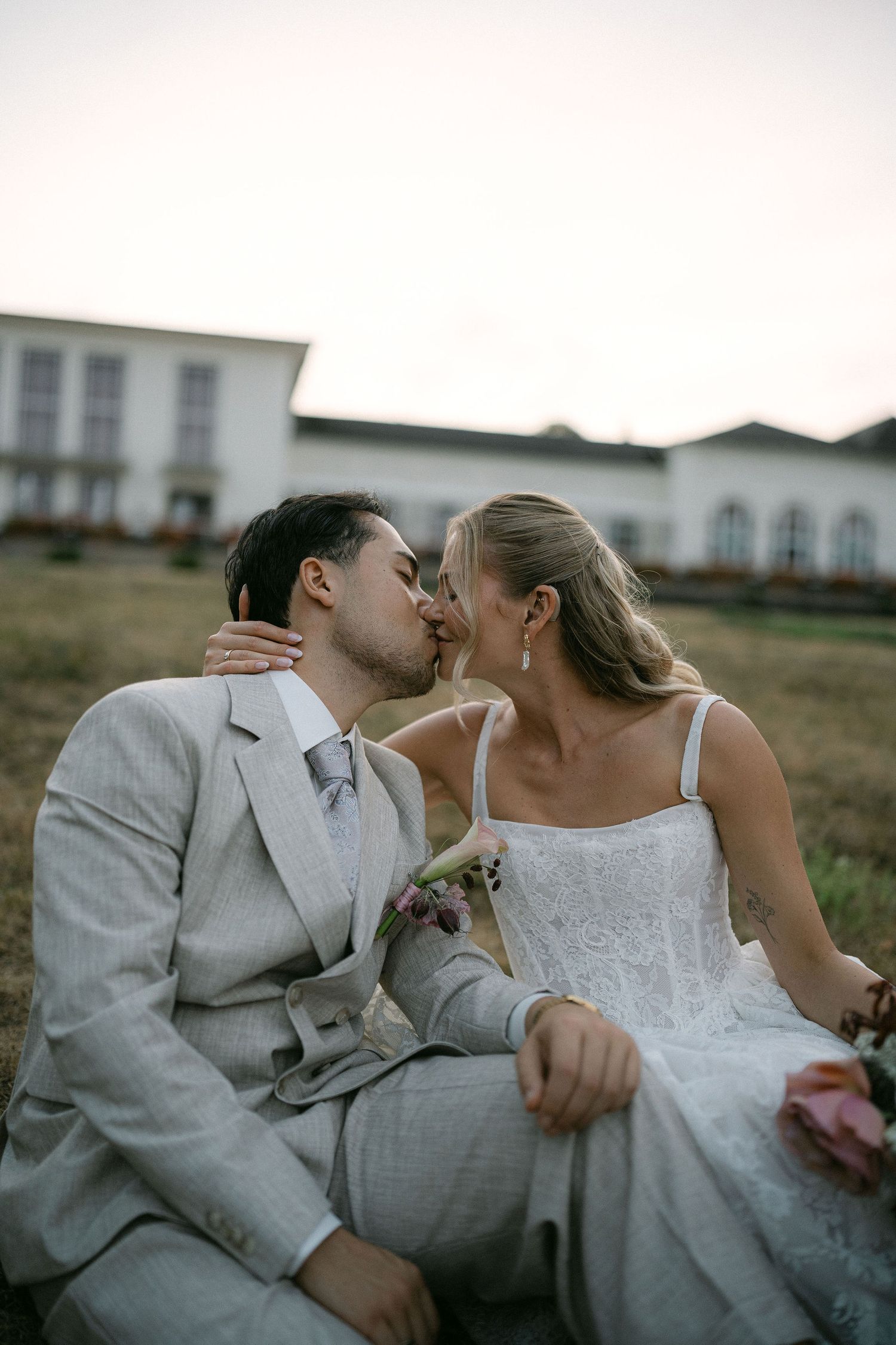Newlyweds share a romantic kiss while sitting on grass at dusk with white wedding venue in the background.