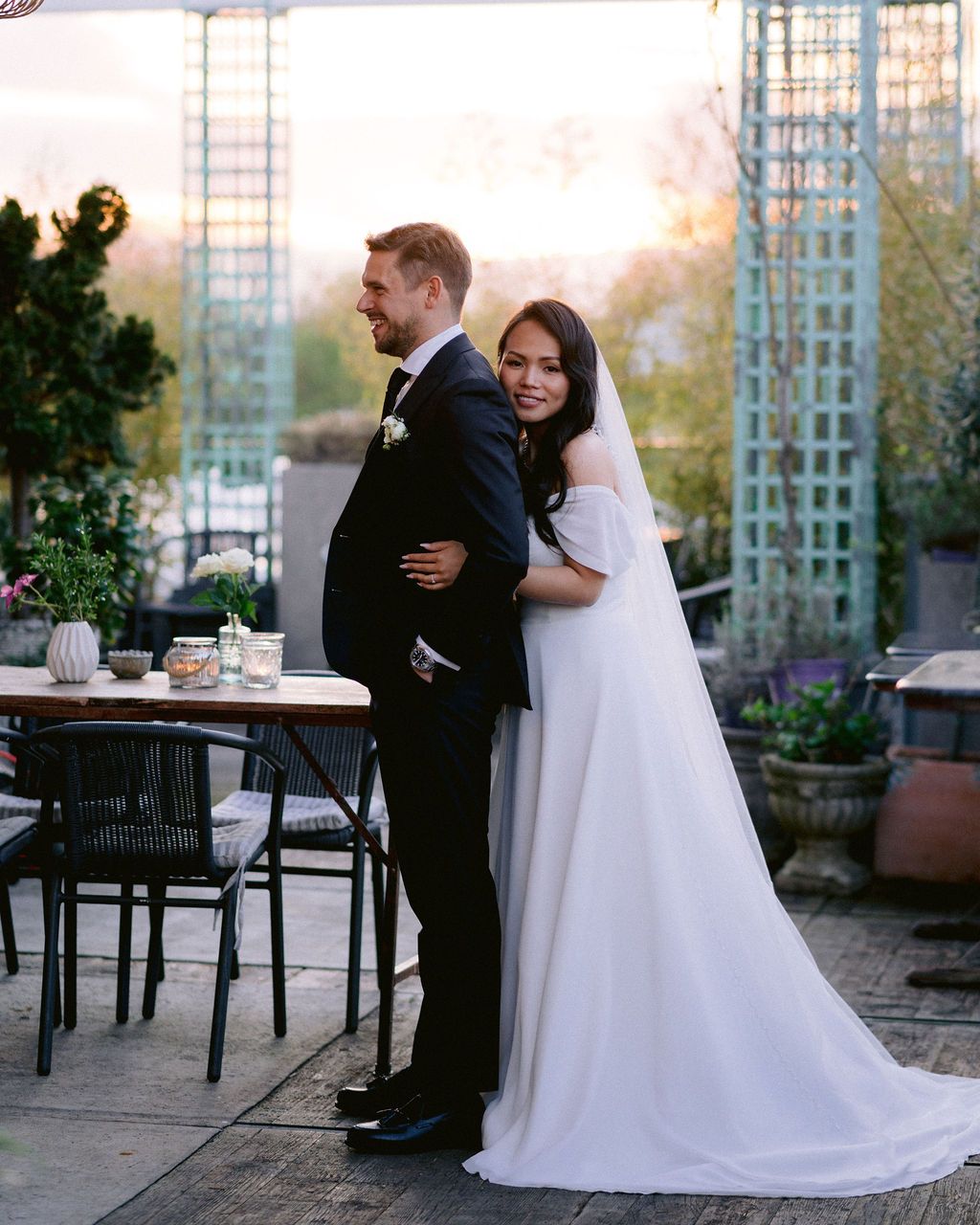 A romantic wedding couple embraces on an outdoor patio at sunset with garden decor and trellis in the background.