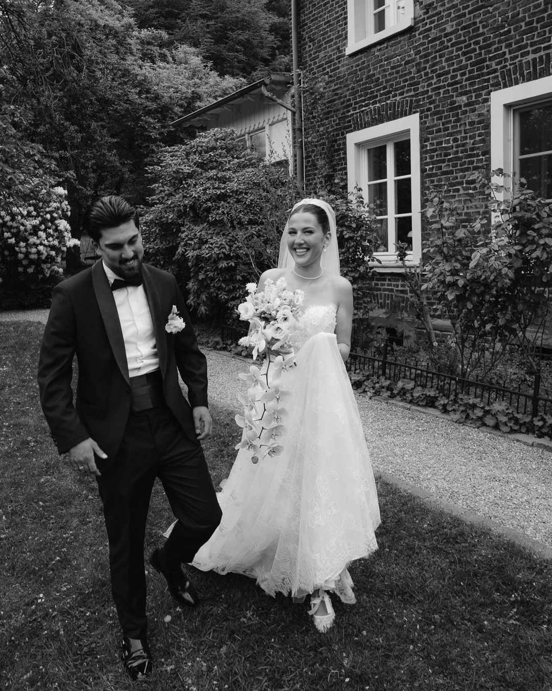 Black and white photo of a smiling bride and groom walking outdoors near a brick building on their wedding day.