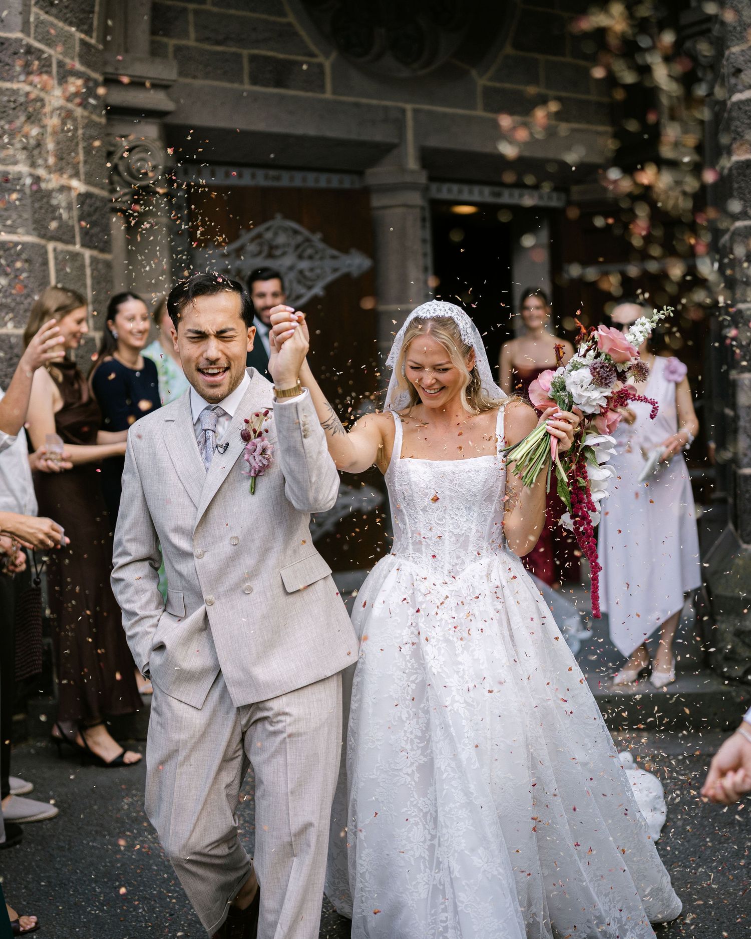 Joyful newlyweds exit church in confetti shower, groom in gray suit, bride in white ballgown with veil, guests celebrating.