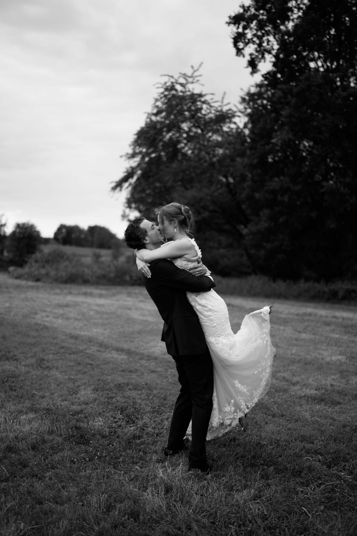 Black and white photo of groom lifting bride in wedding dress outdoors in a field with trees in background.