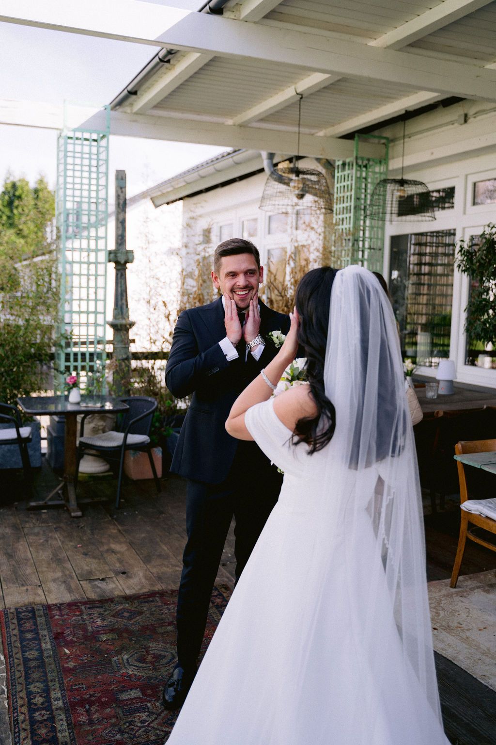 A candid moment between a bride in white wedding gown and veil and groom in dark suit on an outdoor patio.