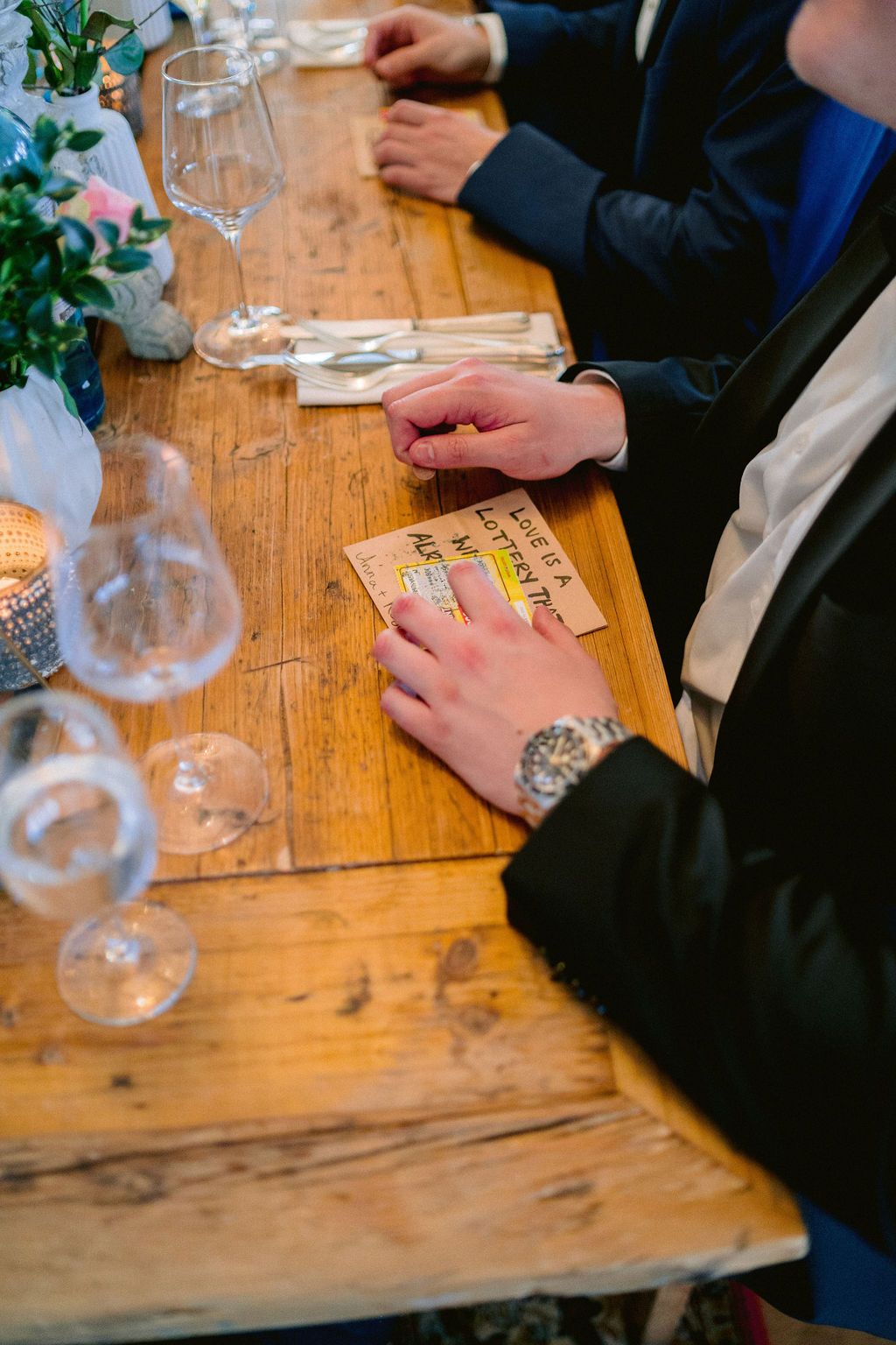 Hands holding playing cards and wine glasses on a rustic wooden table during a game night gathering.