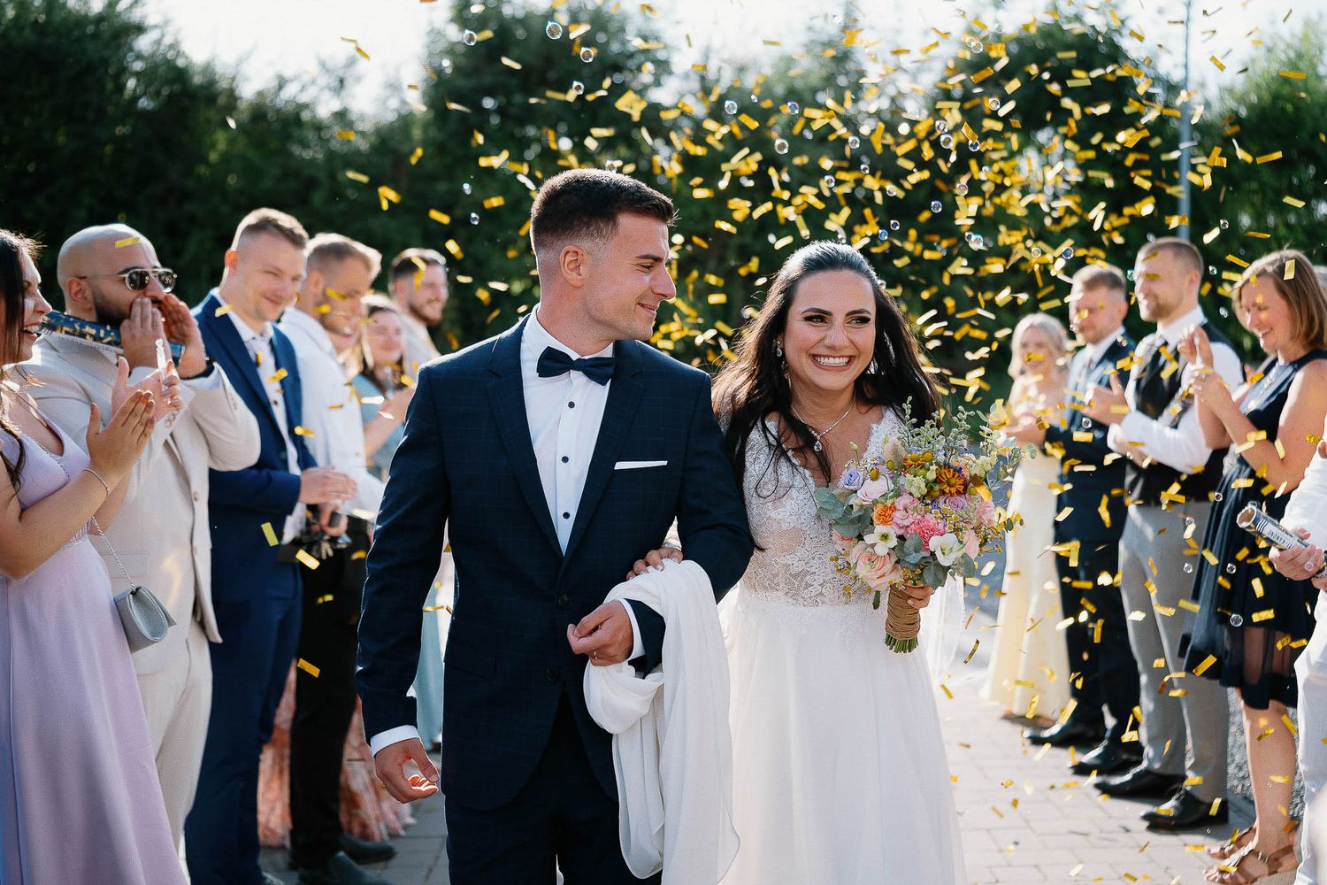Happy couple walks through rows of guests tossing yellow flower petals during outdoor ceremony exit.