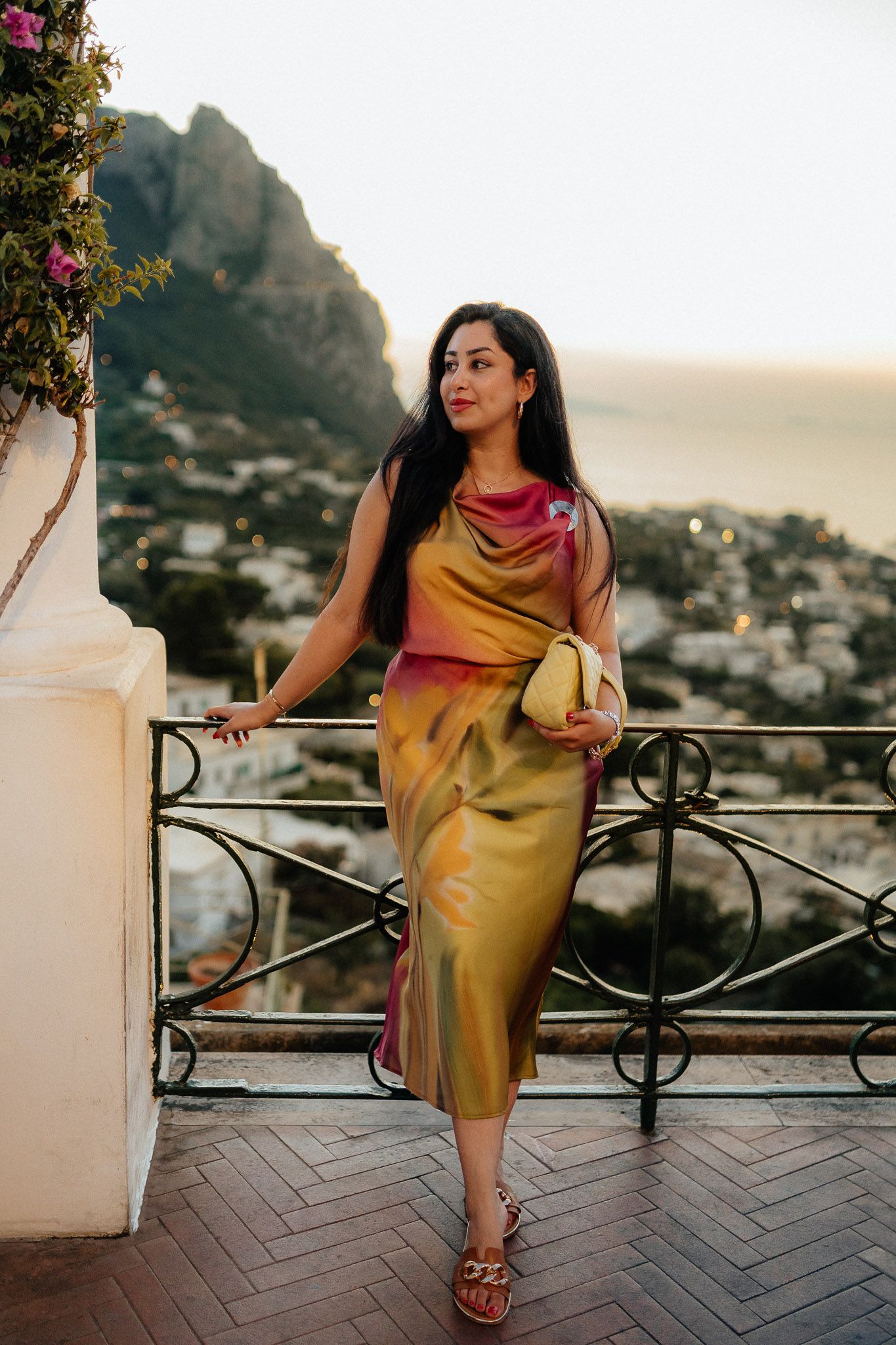 Woman in gold silk midi dress on scenic terrace overlooking coastal town at sunset.