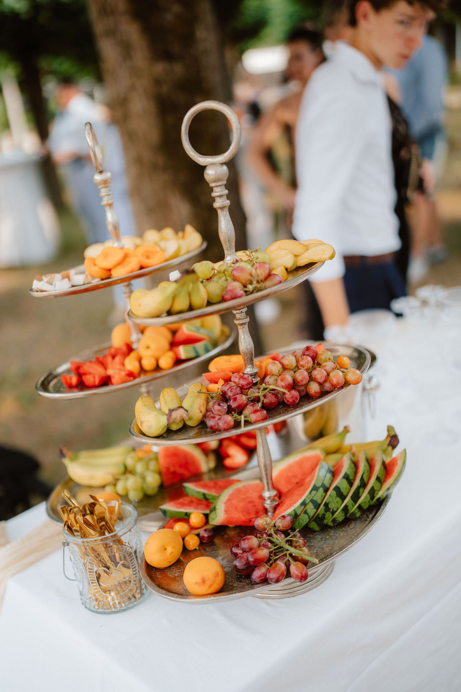 Elegant three-tier silver fruit stand displaying watermelon, grapes, and tropical fruits at an outdoor wedding reception.