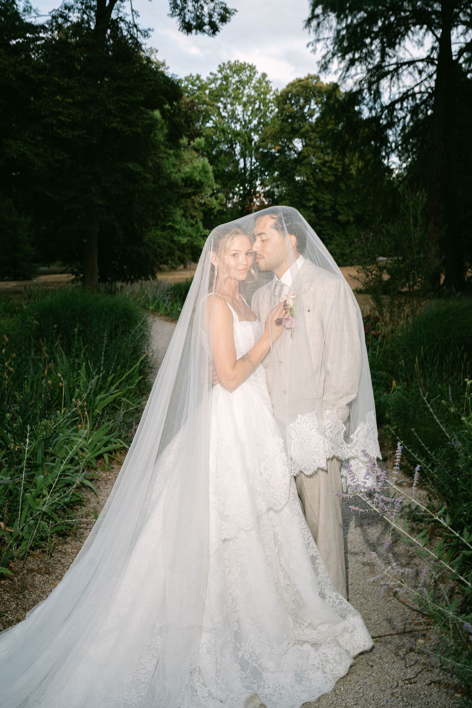 Bride and groom share romantic moment under flowing lace veil in lush garden setting.