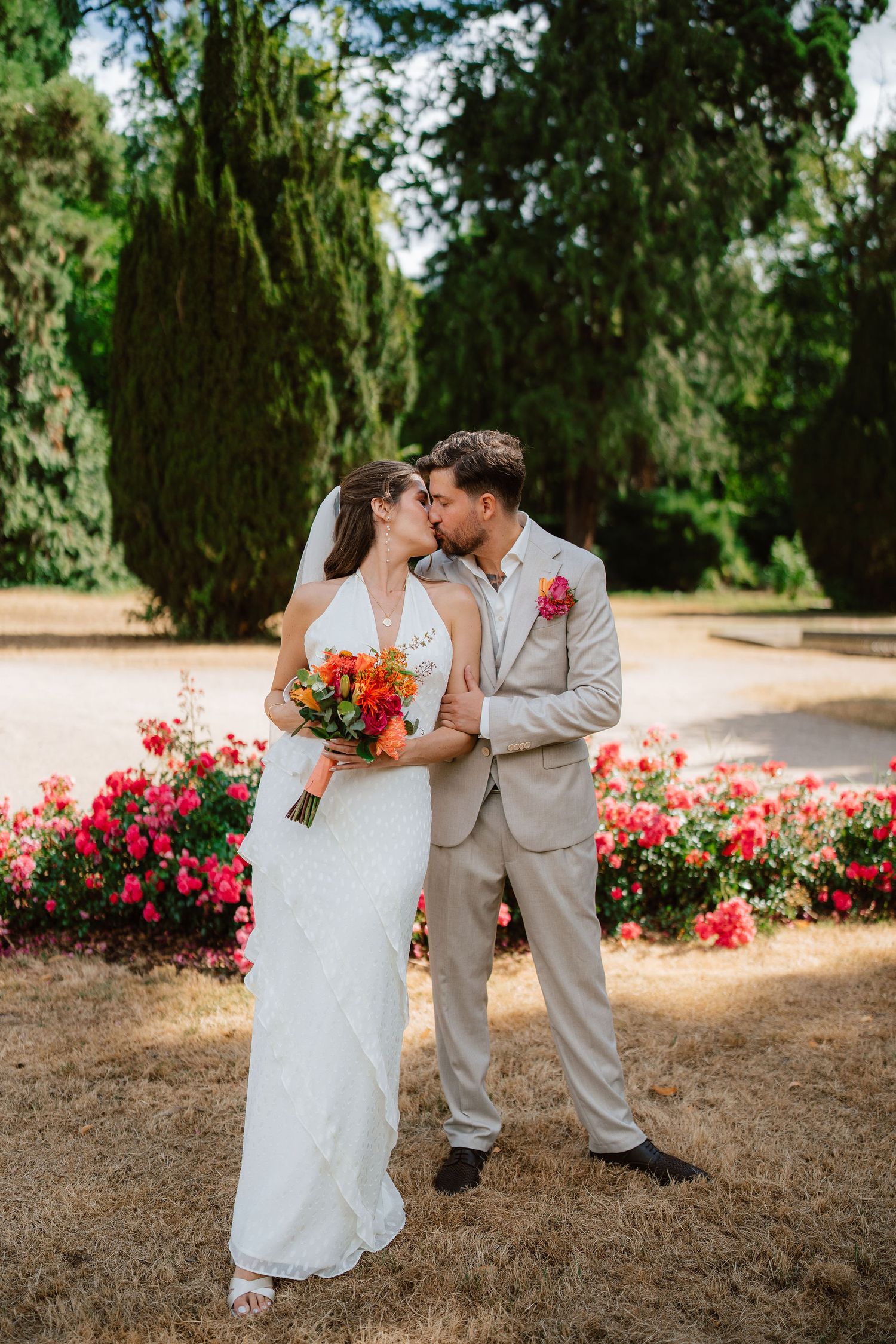 Bride and groom kissing in a garden, surrounded by vibrant red and pink flowers, bride holding a colorful bouquet.