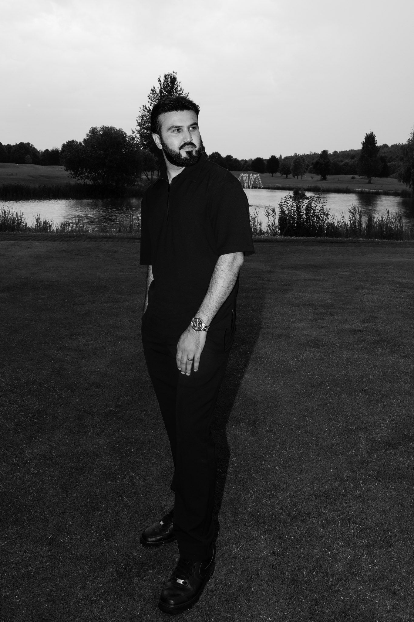 Black and white portrait of person in dark clothing standing by a lake with trees in background at dusk.