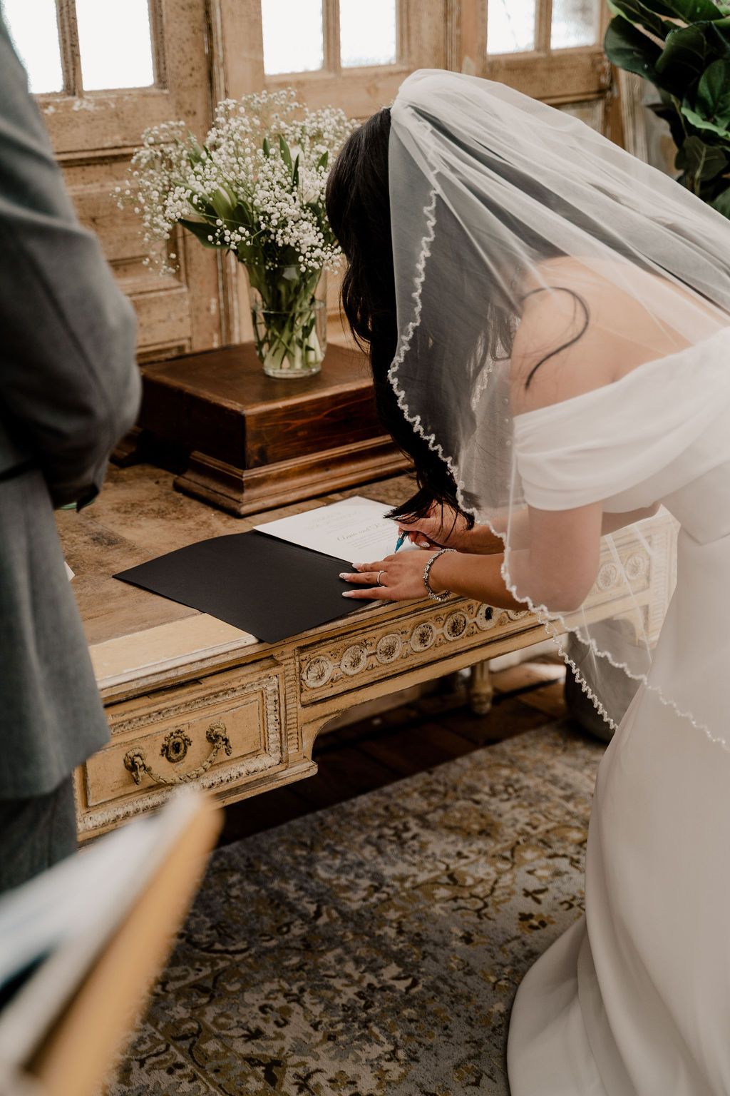 A couple in wedding attire signs the marriage register at a rustic wooden table by a window during their ceremony.