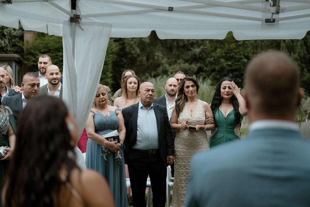 A group gathers under a white tent canopy during an outdoor wedding ceremony.